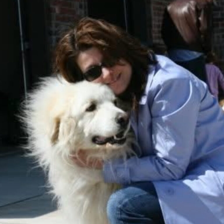 A woman wearing sunglasses and a light blue jacket hugs a fluffy white Great Pyrenees dog outdoors, both appearing happy and content in the sunlight.